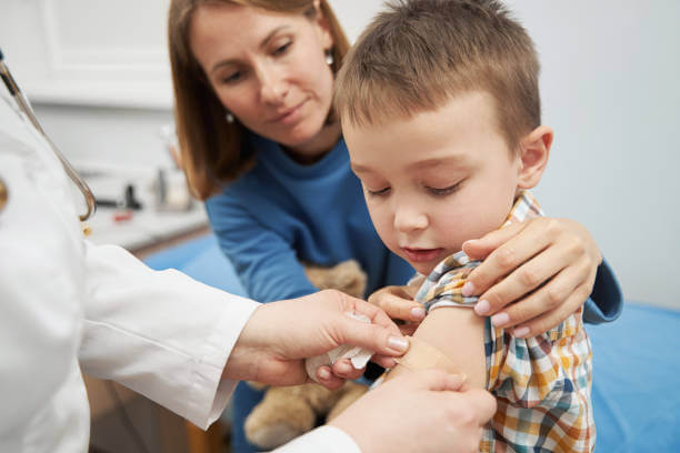 Little boy sitting next to mother and looking at arm while pediatrician putting adhesive bandage on wound after injection