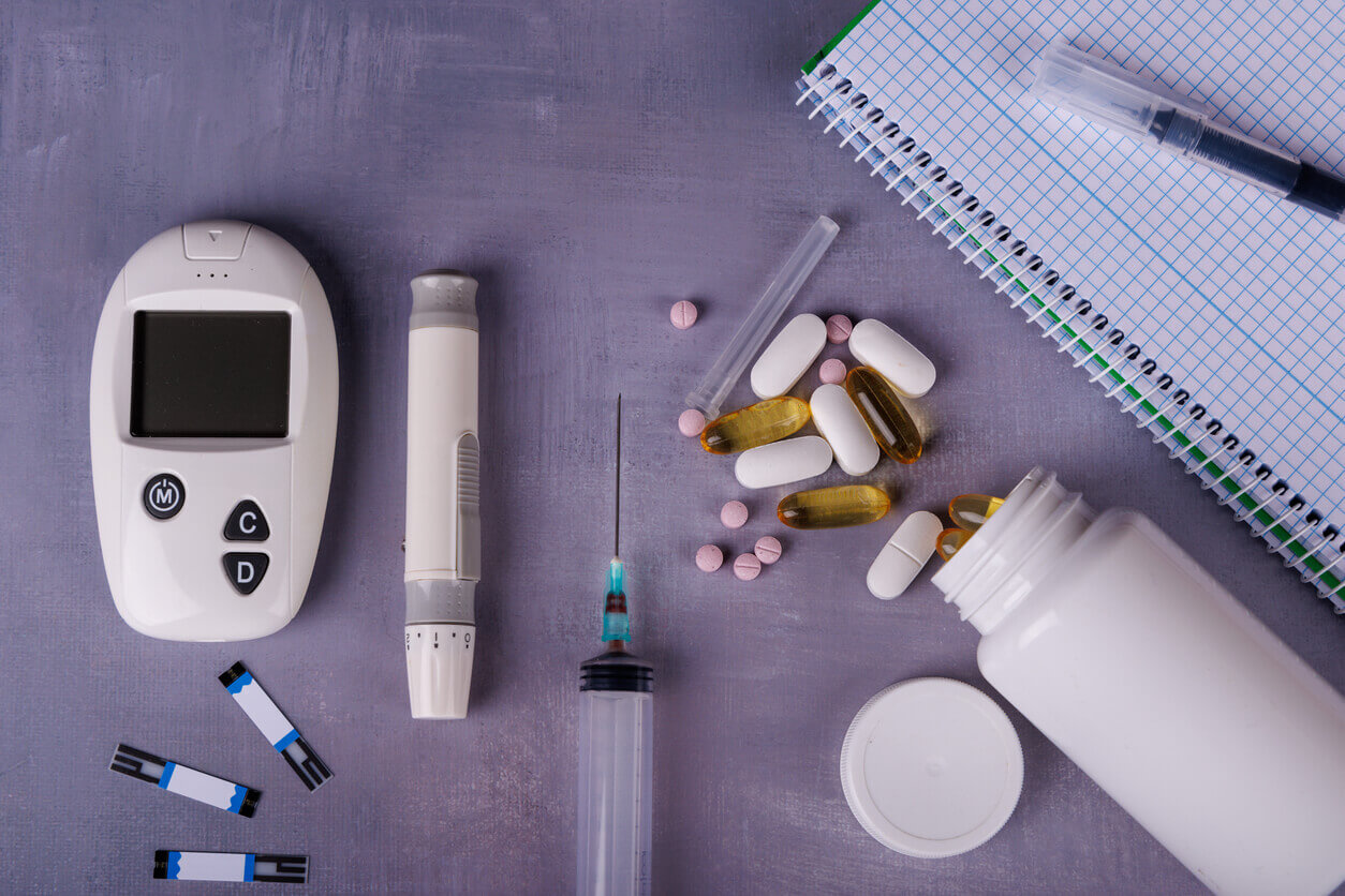 Image of equipment for the medical management of diabetes mellitus, including a glucometer, thermometer, syringe, white pills, and a notebook arranged on a concrete table.